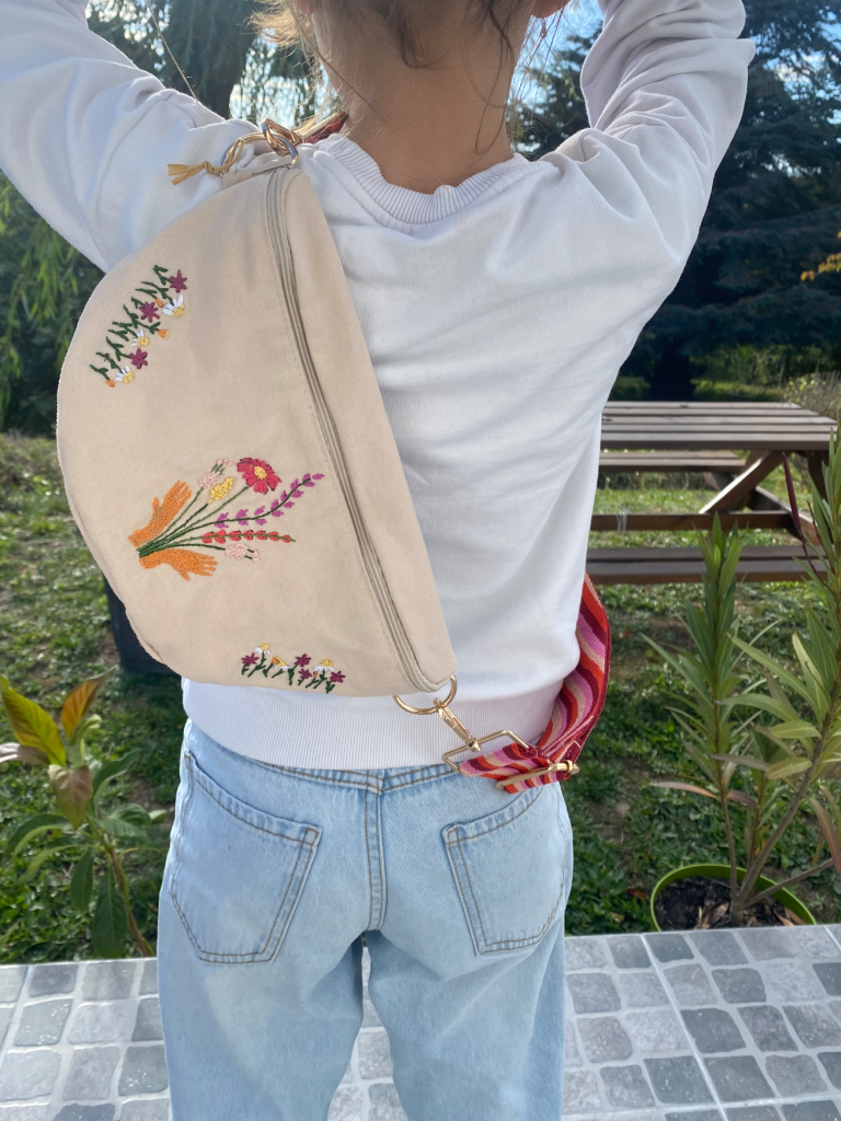 Sac Banane Beige, collection florale marguerites et roses. Sac en toile beige avec broderies florales porté sur l'épaule, devant un paysage verdoyant.